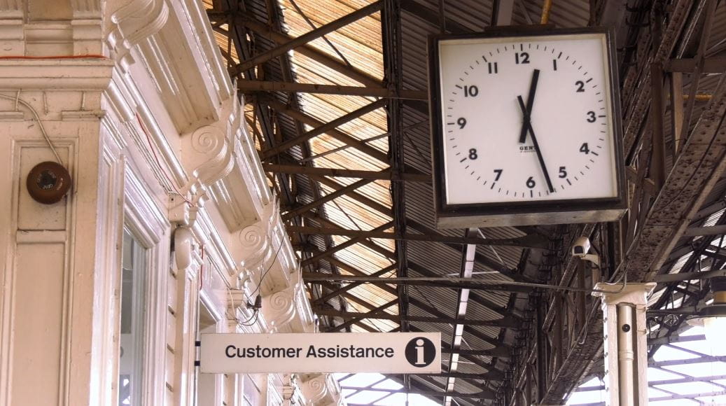 Huddersfield train station with a clock in the foreground and a sign reading Customer Assistance in the background.
