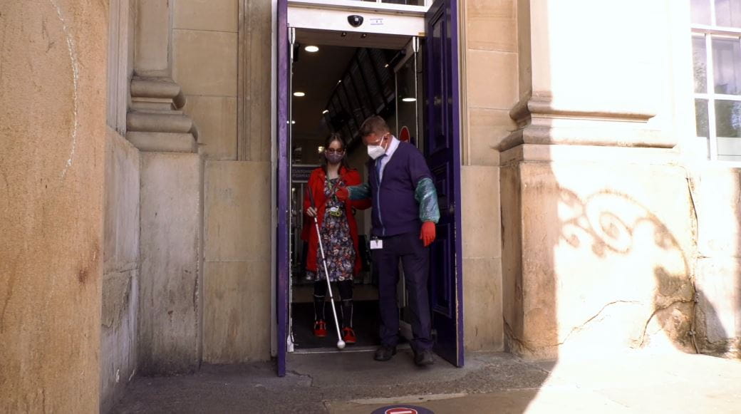 Award-winning disability blogger Chloe Tear receives passenger assistance at Huddersfield train station from a TransPennine Express colleague wearing PPE. They are arm-in-arm walking through the station exit.