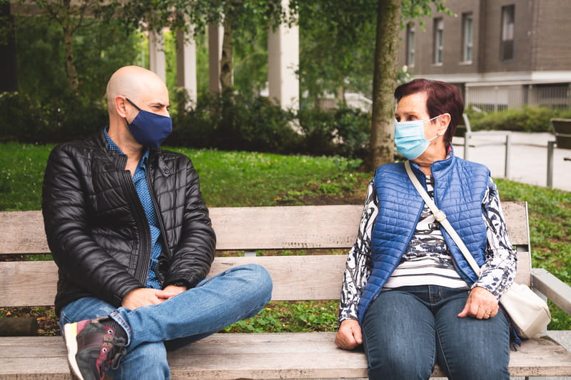 Man wearing face covering talking to elderly woman wearing face covering, both sat on a park bench